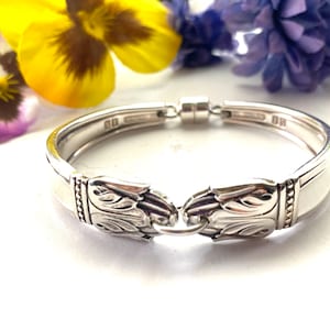 以下が含まれることがあります： 装飾的な花の模様のシルバーブレスレット。ブレスレットは再利用された銀食器で作られており、留め金が付いています。ブレスレットは、黄色と紫の花の背景に対して表示されています。