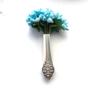 May include: A miniature bouquet of pale blue artificial flowers with green foliage, presented in a silver spoon-shaped vase. The spoon features an elaborate floral pattern on its handle. The arrangement is set against a white backdrop.