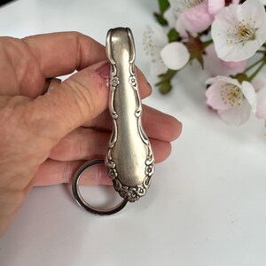 Könnte beinhalten: Schlüsselanhänger aus einem silbernen Löffelstiel. Der Löffelstiel ist mit floralen und scrollartigen Details verziert. Ein silberner Schlüsselring ist befestigt. Im Hintergrund befinden sich weiße und rosa Blüten.
