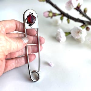 May include: A large silver safety pin with a decorative top. The top features a red oval gemstone set in a silver frame with ornate detailing. The pin is held in a hand, with a white background and floral accents.