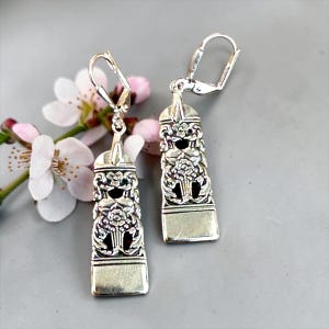 May include: A pair of silver-coloured, rectangular drop earrings with detailed floral and scrollwork patterns. The earrings feature lever-back fastenings and are set against a soft grey backdrop with delicate pink and white flowers.