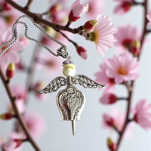 May include: A silver-toned angel pendant necklace with a pearl-like body, detailed wings, and a decorative headpiece. The necklace hangs from a delicate chain, set against a backdrop of pink blossoms.