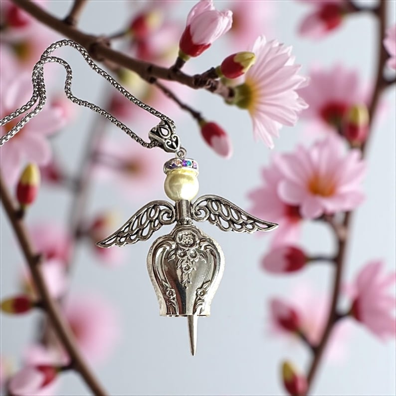 May include: A silver-toned angel pendant necklace with a pearl-like body, detailed wings, and a decorative headpiece. The necklace hangs from a delicate chain, set against a backdrop of pink blossoms.