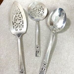 May include: A set of three silver-toned serving utensils including a pointed cake server with a pierced filigree pattern, a round slotted spoon with a central cutout design, and a solid serving spoon, all featuring floral embossed handles.