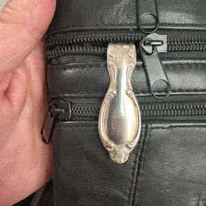 Może przedstawiać: Czarna skórzana torba na ramię z wieloma zamkami błyskawicznymi i ozdobnym srebrnym uchwytem zamka w kształcie łyżki. Torba ma teksturowaną powierzchnię i długi pasek.