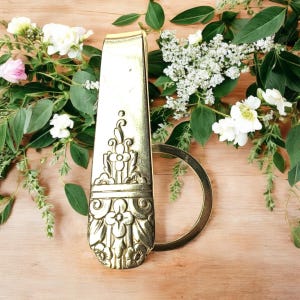May include: A gold-coloured, ornate bottle opener with a floral design. The opener has a long, rectangular handle and a circular ring. The background features white and pink flowers and green foliage on a wooden surface.