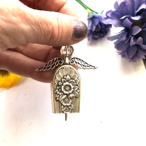 May include: A silver-toned angel figurine with detailed floral embellishments on the body and delicate wing accents. The angel has a crystal head and a small pin at the bottom. The background includes purple flowers.