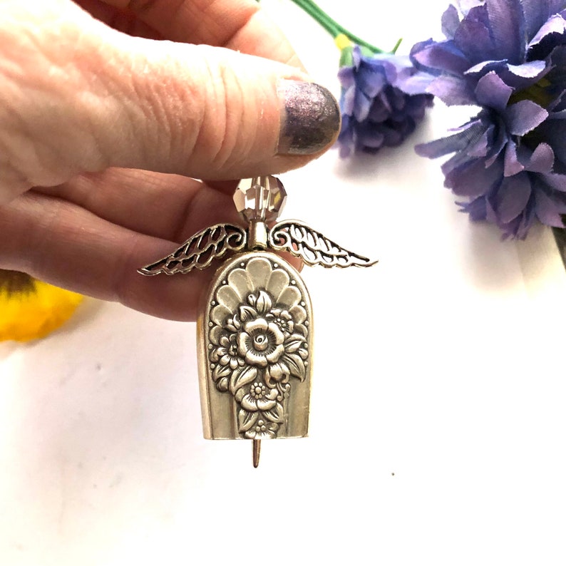 May include: A silver-toned angel figurine with detailed floral embellishments on the body and delicate wing accents. The angel has a crystal head and a small pin at the bottom. The background includes purple flowers.
