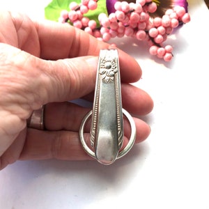 Puede incluir: Un llavero de cuchara de plata con un diseño floral y un anillo de plata unido al mango.