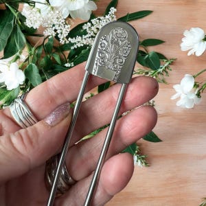 Może przedstawiać: Srebrna szpilka do włosów z ozdobnym, kwiatowym wzorem na dekoracyjnej główce. Szpilka jest prezentowana na tle białych kwiatów i zieleni, podkreślając jej szczegóły i styl.