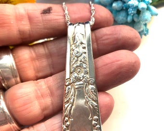 Puede incluir: Un collar de plata con un colgante hecho de una pieza de cubiertos vintage. El colgante presenta un diseño floral y está unido a una cadena delicada.