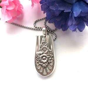 May include: A silver necklace with a pendant shaped like a spoon with a floral design. The pendant is detailed with a large flower and smaller flowers around it.