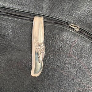 Puede incluir: Un tirador de cremallera plateado con detalles florales está sujeto a una bolsa de cuero negro. La cremallera está cerrada y el tirador es un elemento decorativo. El cuero tiene un aspecto texturizado.
