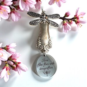 May include: A silver dragonfly ornament made from a spoon, with a circular charm that reads "Now She flies with dragonflies." Pink blossoms frame the ornament against a white background.