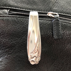 May include: A silver-toned metal zipper pull with a floral design. The pull is attached to a black leather bag.