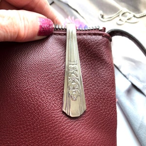 Puede incluir: Un bolso de cuero burdeos con un tirador de cremallera plateado hecho de un tenedor vintage. El mango del tenedor presenta un diseño floral en relieve. El bolso tiene una correa oscura y una superficie texturizada.