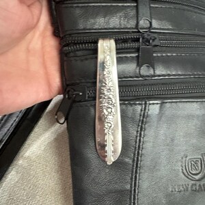 Puede incluir: Un bolso bandolera de cuero negro con múltiples compartimentos con cremallera. Una cuchara decorativa plateada con detalles florales está sujeta a la cremallera del bolso. El bolso tiene una correa para el hombro y un logotipo en la parte delantera.