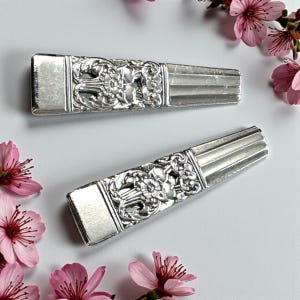 Pode incluir: Dois descansos de faca antigos prateados com desenhos florais e de rolagem ornamentados. Os descansos de faca têm formato retangular com uma seção central detalhada e uma extremidade lisa. Flores cor-de-rosa dispostas ao redor.