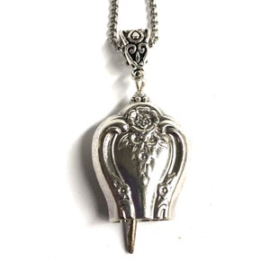 May include: A silver necklace with a bell pendant featuring a floral design. The bell is attached to a silver chain.