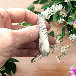 Könnte beinhalten: Ein silberner Löffelring mit floralen Details. Der Ring besteht aus einem Vintage-Löffel und weist zarte geprägte Blumen und Blätter auf. Der Ring wird vor einem Hintergrund aus weißen Blumen und Grün gehalten.