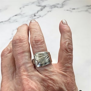 Puede incluir: Un anillo de plata con un diseño de hojas. El anillo es rectangular con un patrón de hojas detallado en la parte superior. El anillo se lleva en un dedo.