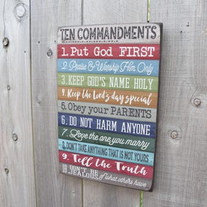 May include: A wooden sign with the Ten Commandments written on it. The sign is painted in a rainbow of colors and has a distressed look. The text on the sign reads: "TEN COMMANDMENTS 1. Put God FIRST 2. Praise & Worship Him Only 3. KEEP GOD'S NAME HOLY 4. Keep the Lord's day special 5. Obey your PARENTS 6. DO NOT HARM ANYONE 7. Love the one you marry 8. DON'T TAKE ANYTHING THAT IS NOT YOURS 9. Tell the Truth 10. DON'T BE JEALOUS of what others have."
