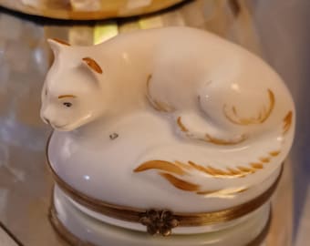 May include: A white porcelain trinket box shaped like a cat lying down. The cat has gold accents and a gold flower on the lid.