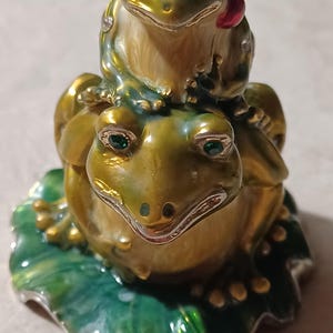 May include: A gold and green frog figurine with a smaller frog figurine on top. The frogs are sitting on a green leaf.