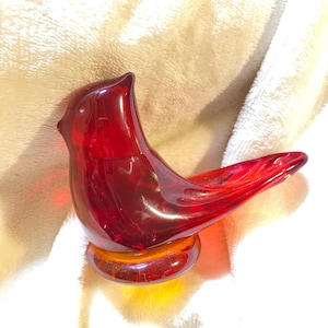 May include: A vibrant red glass bird figurine with a rounded body and a long, pointed tail. The bird is perched on a circular, amber-colored base. The figurine is made of transparent glass, allowing light to pass through.