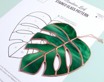 May include: A stained glass Monstera leaf in shades of green with copper-colored edging, lying on a pattern sheet. The pattern sheet has a line drawing of the leaf and the text "Monstera Leaf STAINED GLASS PATTERN".