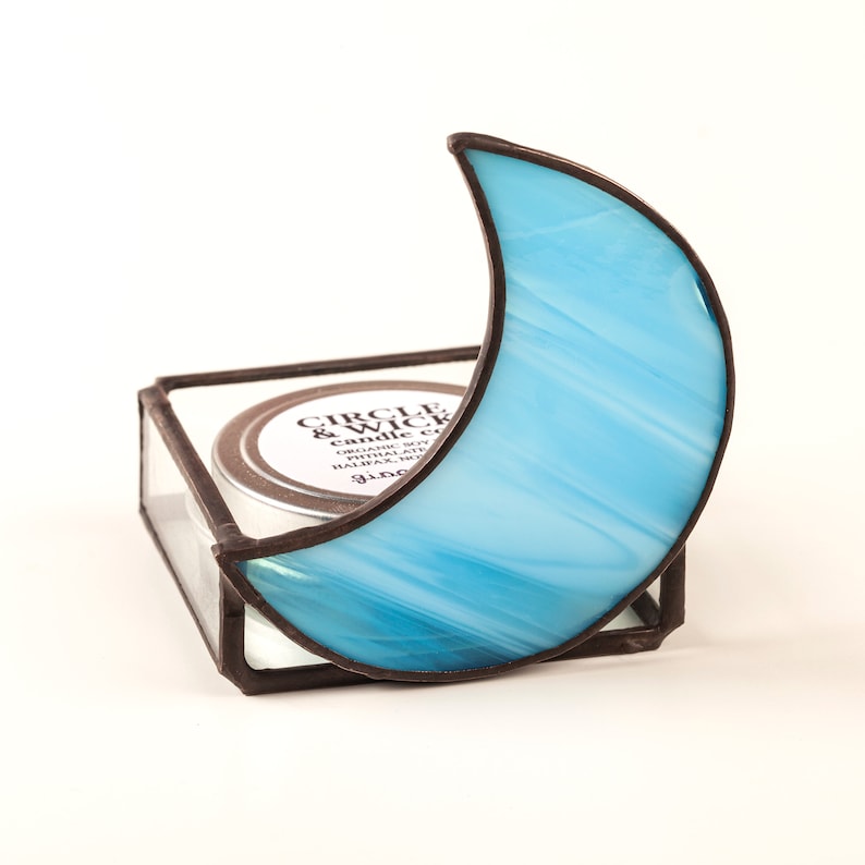 May include: A blue glass crescent moon candle holder with a small, round candle inside. The candle holder is made of stained glass and has a black metal frame. The candle is labeled "Circle & Wick Candle" and is made with organic soy wax, phthalate-free fragrance, and hand-poured in Halifax, Nova Scotia.
