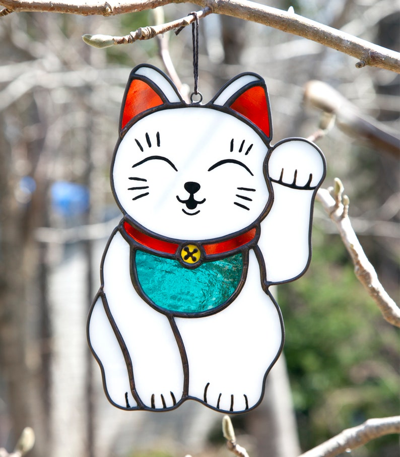 A stained glass lucky cat hanging outdoors in a tree and shows the sun shining through.