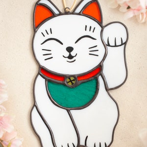 A stained glass lucky cat laying on a table with cherry blossoms surrounding. The cat is white with a red collar with a bell and a green bib. The face details are painted.