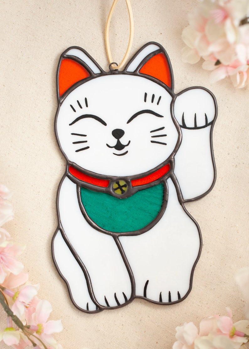 A stained glass lucky cat laying on a table with cherry blossoms surrounding. The cat is white with a red collar with a bell and a green bib. The face details are painted.