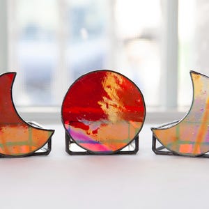 May include: Three stained glass moon and sun shapes in red, orange, and iridescent colors. The shapes are on black metal stands.