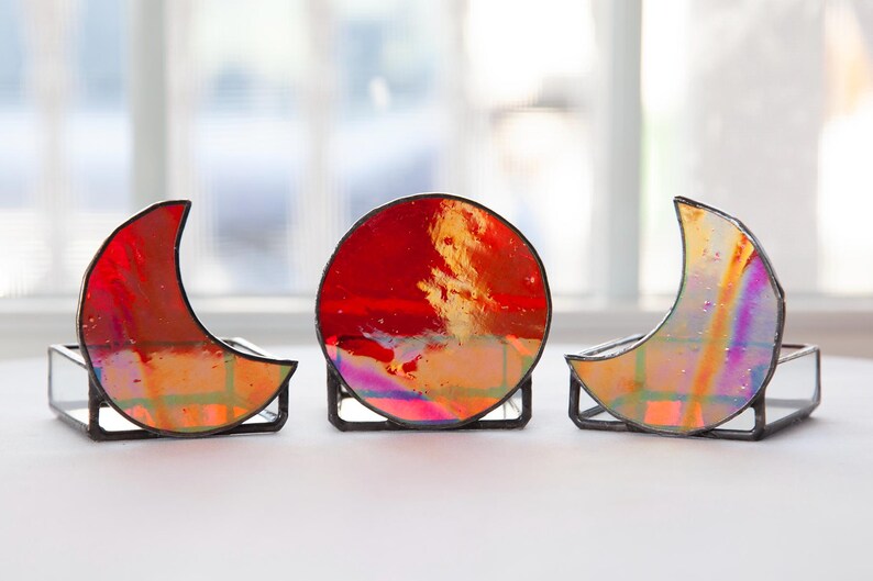 May include: Three stained glass moon and sun shapes in red, orange, and iridescent colors. The shapes are on black metal stands.