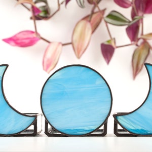 May include: Three stained glass moon and circle candle holders in a light blue color. The candle holders are made of glass and have a black metal frame.