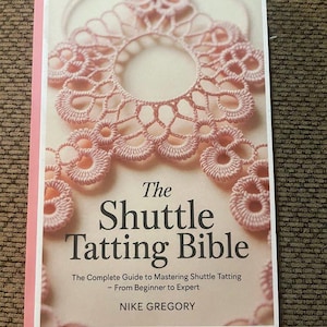 May include: A softcover book titled The Shuttle Tatting Bible by Nike Gregory. The cover features a light pink, intricate lace tatting design against a pale background. The subtitle reads The Complete Guide to Mastering Shuttle Tatting.