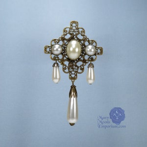 May include: A gold filigree brooch with a large faux pearl in the center and three smaller faux pearls hanging from the bottom. The brooch is in the shape of a cross.