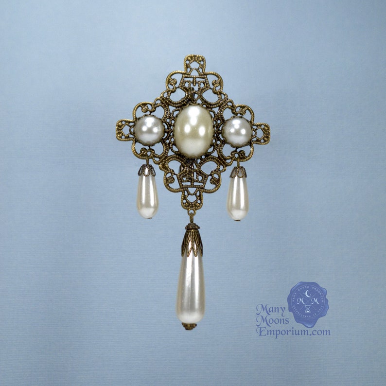 May include: A gold filigree brooch with a large faux pearl in the center and three smaller faux pearls hanging from the bottom. The brooch is in the shape of a cross.