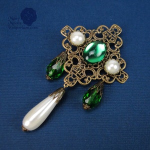May include: A gold filigree pendant with green and white glass beads. The pendant features a square design with intricate details and three dangling beads.