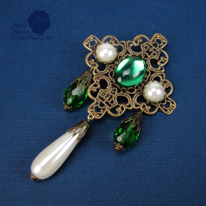 May include: A gold filigree pendant with green and white glass beads. The pendant features a square design with intricate details and three dangling beads.