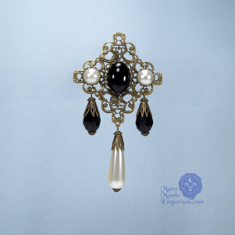 May include: A gold filigree brooch with black and white faux pearls. The brooch features a black oval stone in the center and three dangling pear-shaped stones.