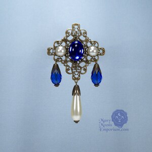 May include: A gold filigree brooch with a blue gemstone and white pearl accents. The brooch has a dangling pearl drop.