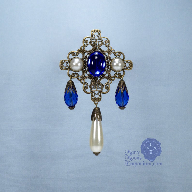 May include: A gold filigree brooch with a blue gemstone and white pearl accents. The brooch has a dangling pearl drop.