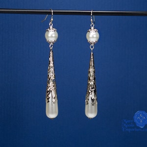 May include: A pair of silver dangle earrings with white pearl accents. The earrings feature a teardrop-shaped design with intricate filigree details.