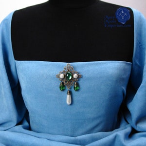 May include: A silver filigree brooch with green and white gemstones. The brooch is on a light blue fabric background.