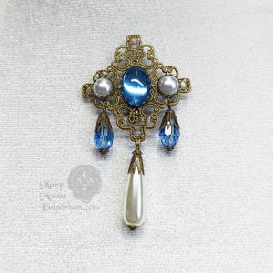 May include: A gold filigree brooch with a blue gemstone and white pearl accents. The brooch has a dangling pearl drop.