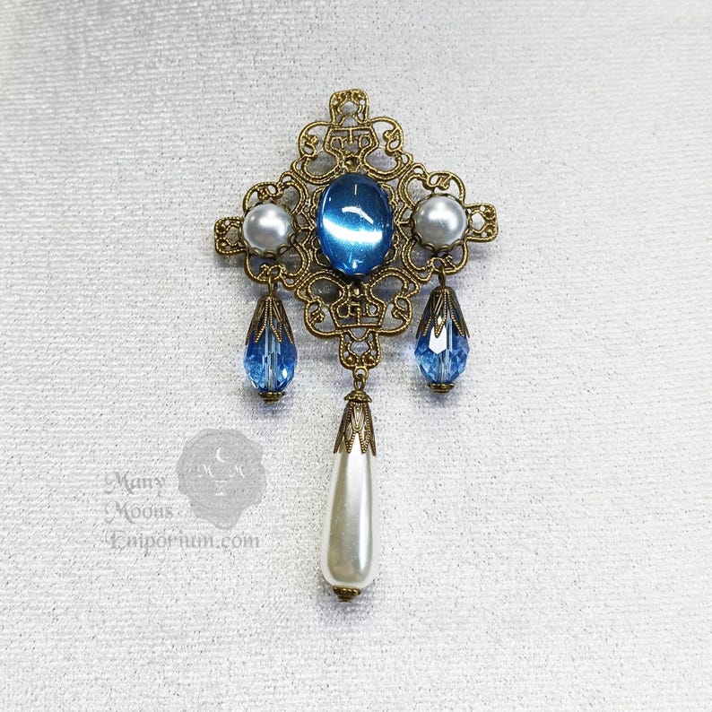 May include: A gold filigree brooch with a blue gemstone and white pearl accents. The brooch has a dangling pearl drop.