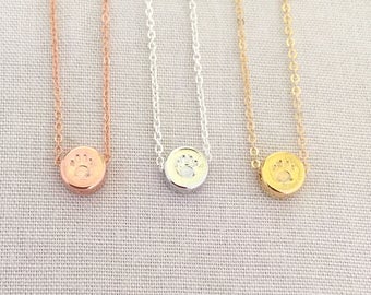 May include: Three delicate necklaces with a small, round pendant featuring a paw print design. The necklaces are available in rose gold, silver, and gold.
